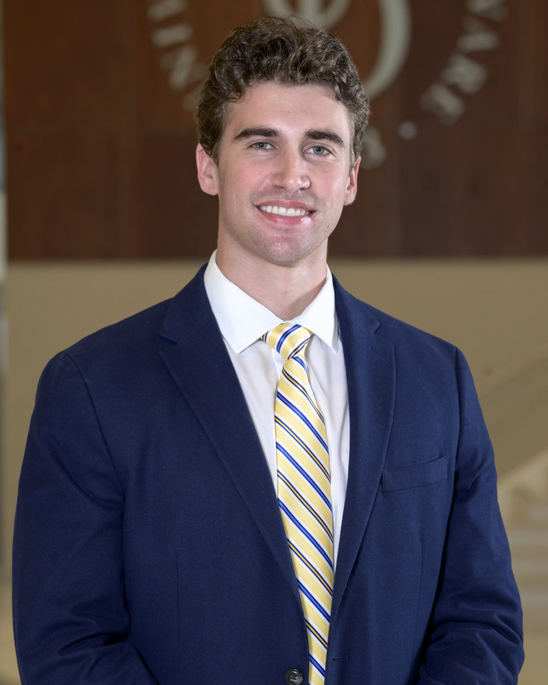 A headshot of Grant Maxa in a suit and tie, with a UD logo behind him. 