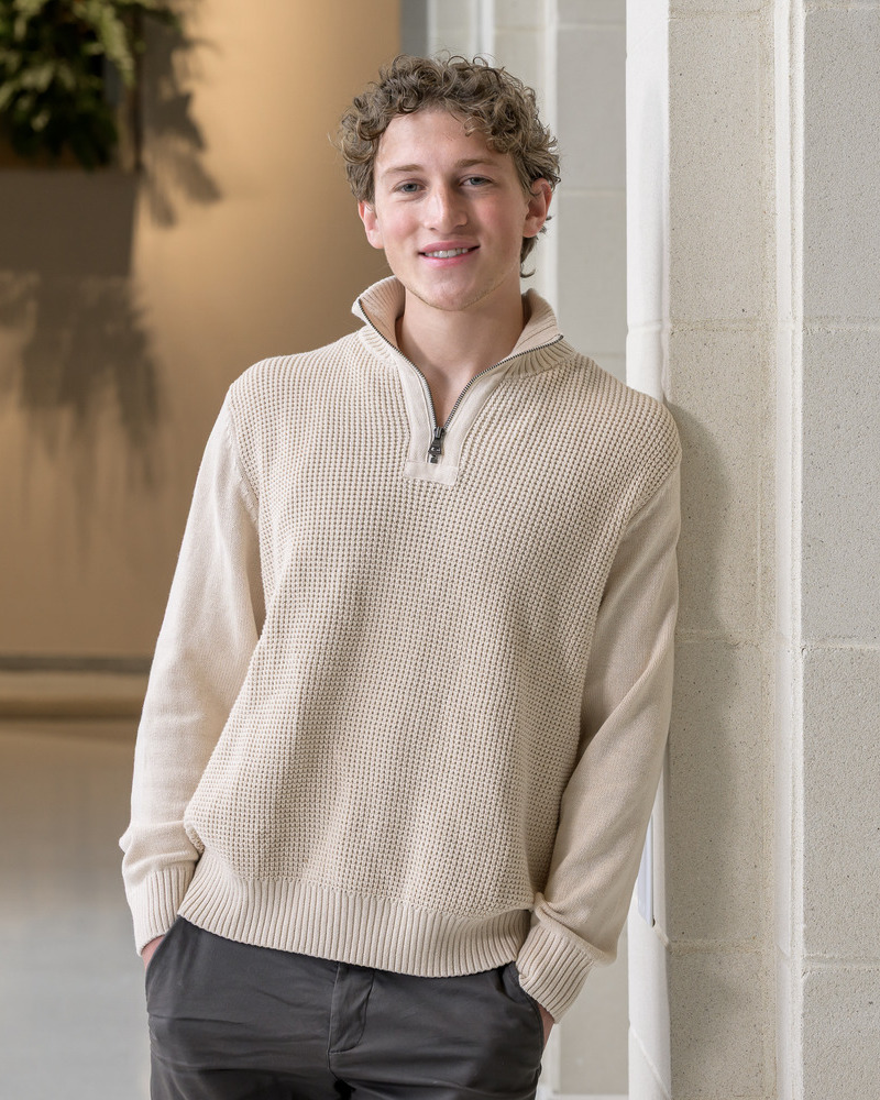 A head of senior Leonard Rein, a human physiology major, standing against a wall with his hands in his pockets. He's wearing a beige collared weater and brown pants.