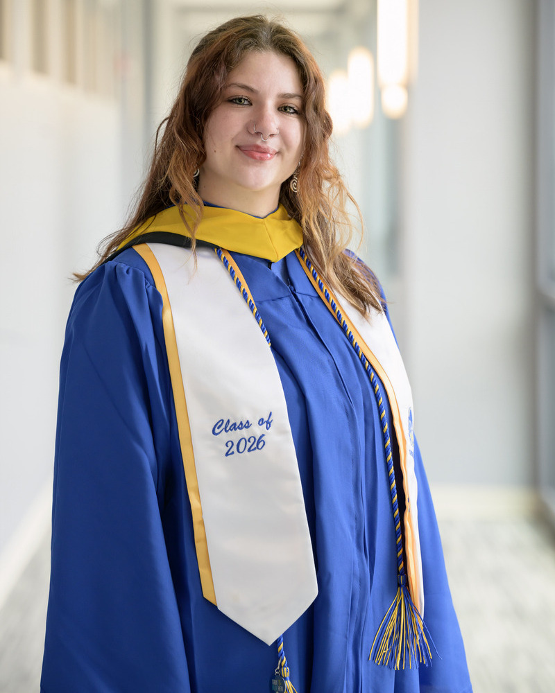 A headshot of senior human phyisology major Emma Rudolph standing to the side wearing her blue and gold offical Commencement regalia