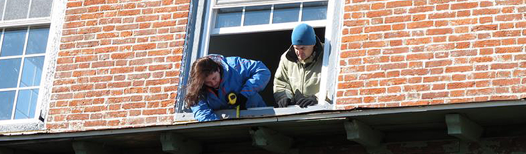 View from below looking up at two students as they lean out of a window and work on restoring a brick building.