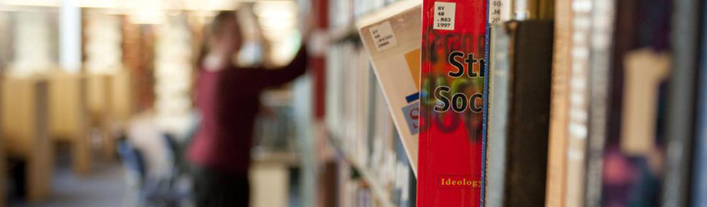 An in-focus book partly pulled out of a library shelf with an out of focus woman in the background.