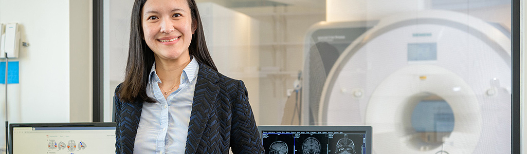A researchers smiles in front of an MRI with brain scans on the computer behind her.