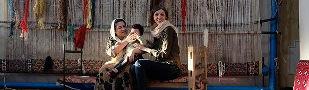 A doctoral student poses with a Kurdish woman and baby with textiles for weaving hanging in the background.