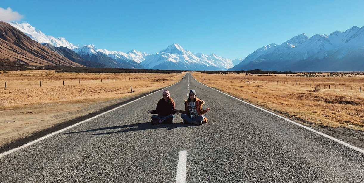 A photo of two students sitting along a road against a backdrop of New Zealand Mountains.