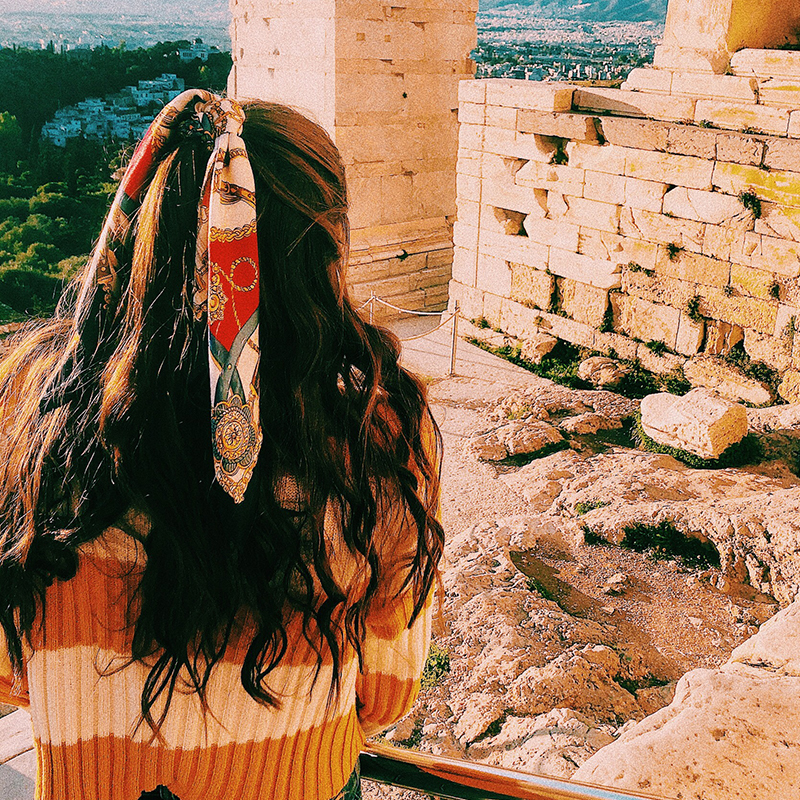 A girl stands overlooking an ancient Greek structure. She has a ribbon in her hair.