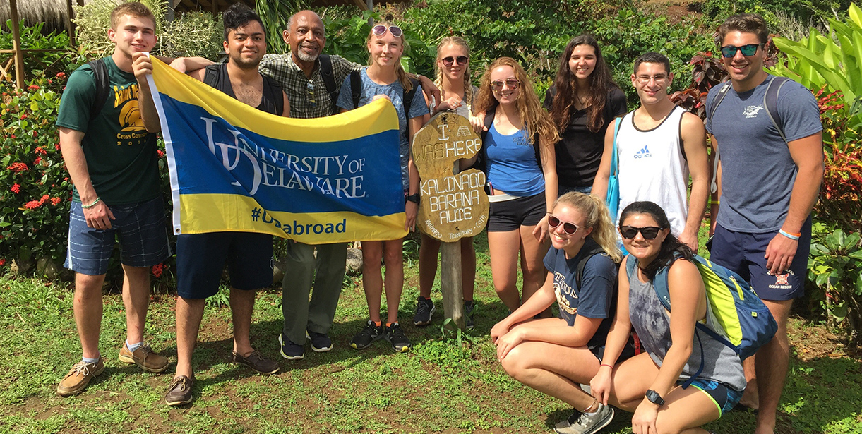 A group of students and their Study Abroad Faculty Director stand for a photo with the UD Flag in Dominica.