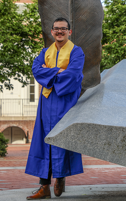 UD World Scholar Jed Hackett poses for a photo in his grad regalia by the UD Book Statue.
