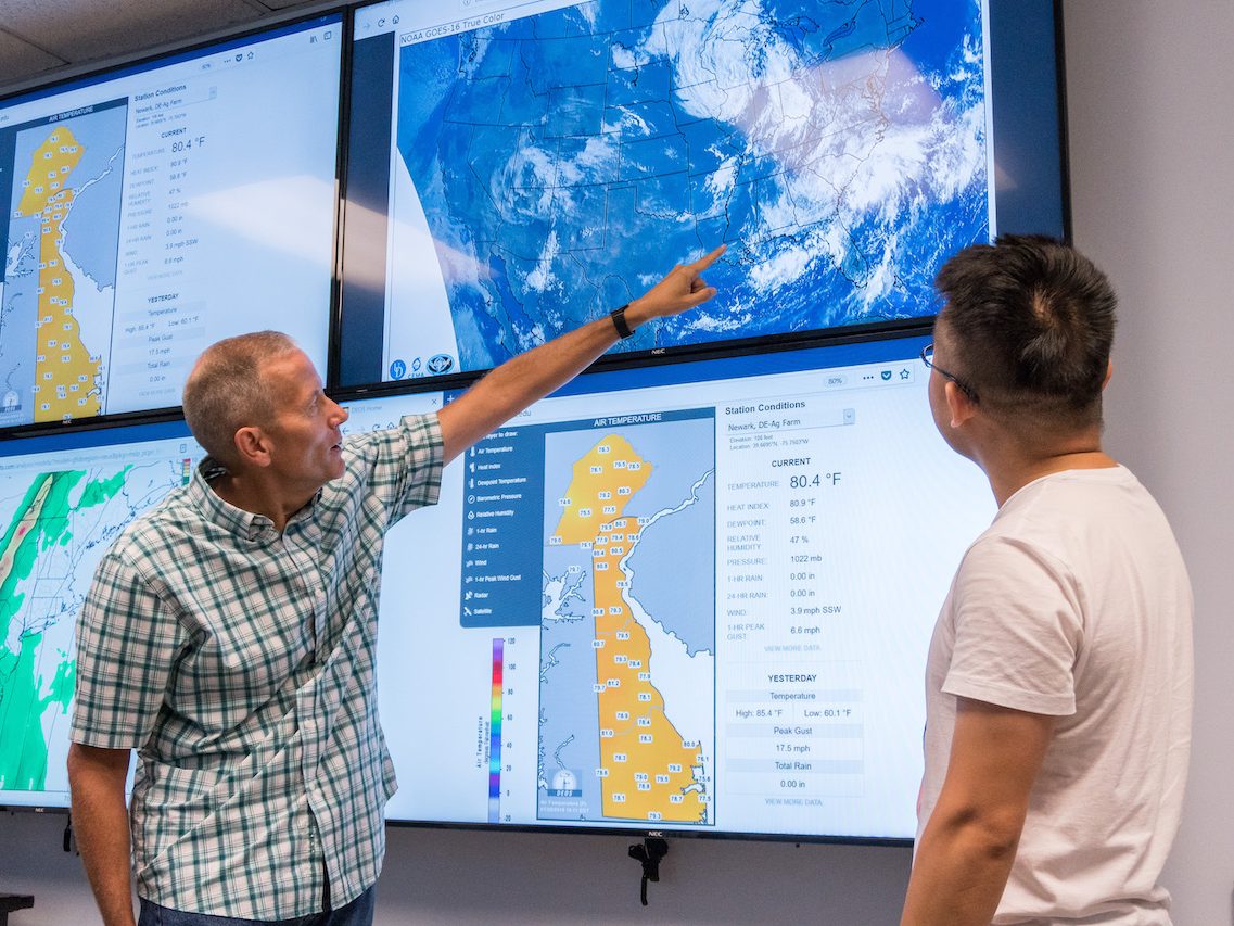 Two men look at digital screens that display weather data and a map of Delaware.