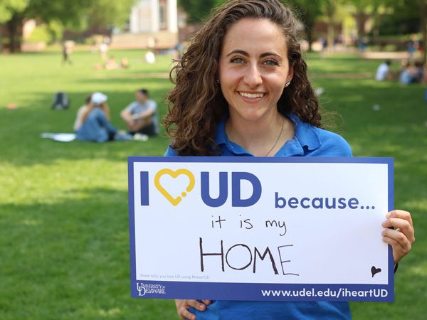 Young woman holds an "I Heart UD because it is my home" sign while smiling on the green.