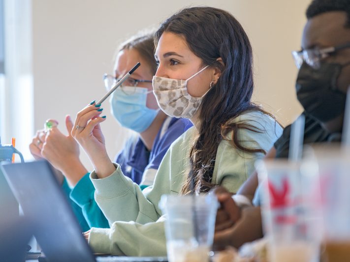 Three students in masks sit and take notes during a class. Three students in masks sit and take notes during a class.