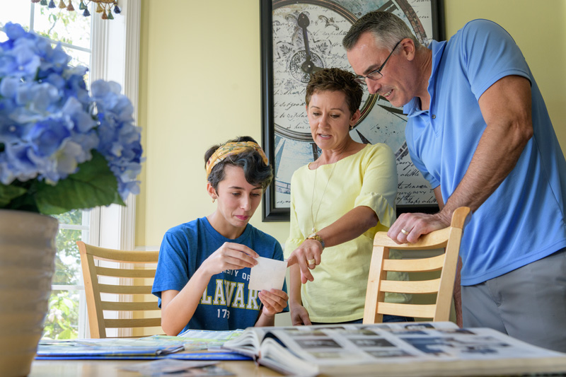 A UD student sits at a table while her parents stand over her looking at photographs together. 