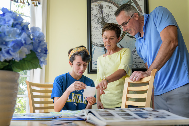 A UD student sits at a table while her parents stand over her looking at photographs together. 