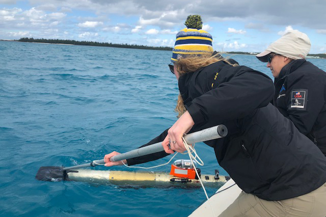 Two UD researchers stand on the edge of a boat and use a long pole to hook at research vessel in the ocean. 