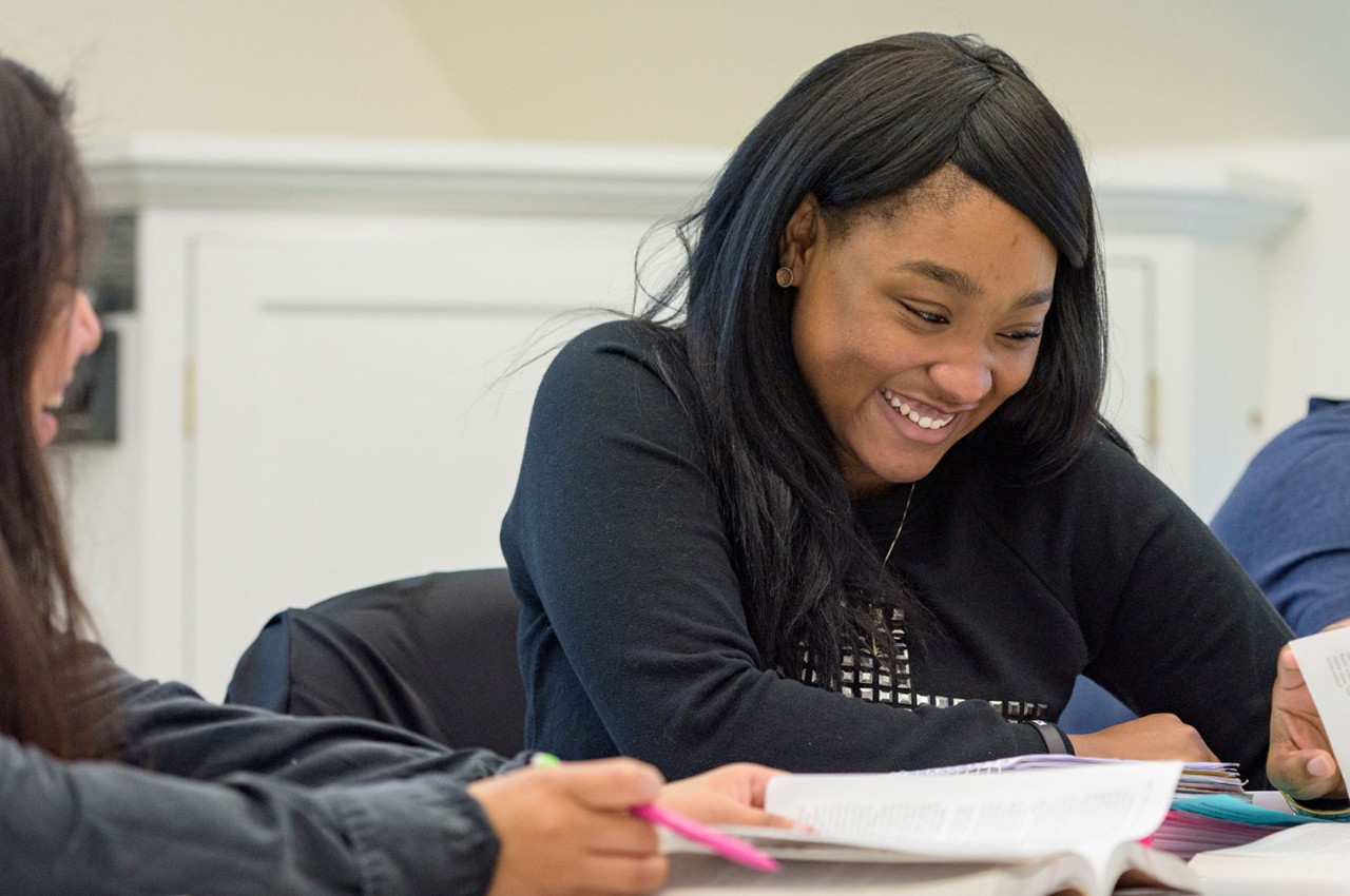 Image of two young women smiling as they look down at work on a desk. 