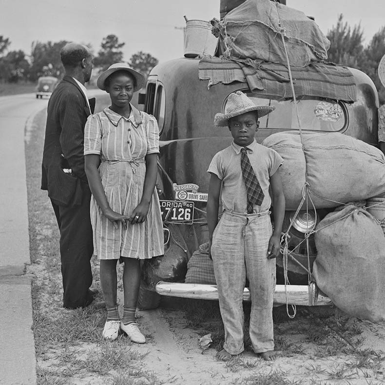A Black and white photo of a family standing on the side of the road by their packed car. 