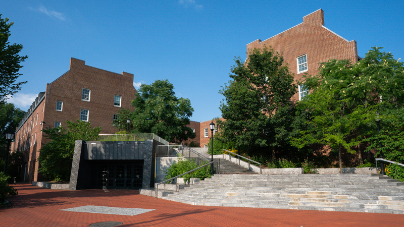 View of the front entrance of the Caesar Rodney complex. There are large stairs in the middle of the picture that go up to a second-story entrance, with the Caesar Rodney buildings behind the staircase. To the left of the stairs is the entrance to the Caesar Rodeny Dining Hall, and there is a brick walkway leading up to the stairs and dining hall entrance. 