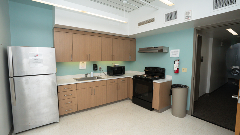 A refrigerator sits in the left corner, with cabinets next to it along the back wall. There is a sink in the countertop and also a microwave sitting on top. The oven and stove is on the right wall next to the entry/exit door, with a trashcan and also fire extenguisher mounted on the wall.y complex. There are large stairs in the middle of the picture that go up to a second-story entrance, with the Caesar Rodney buildings behind the staircase. To the left of the stairs is the entrance to the Caesar Rodeny Dining Hall, and there is a brick walkway leading up to the stairs and dining hall entrance. 