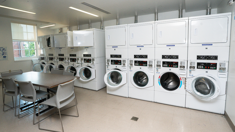 Nine units of washing machines on the bottom and dryers stacked on top along the back wall. A table with chairs around it is in the middle of the room.