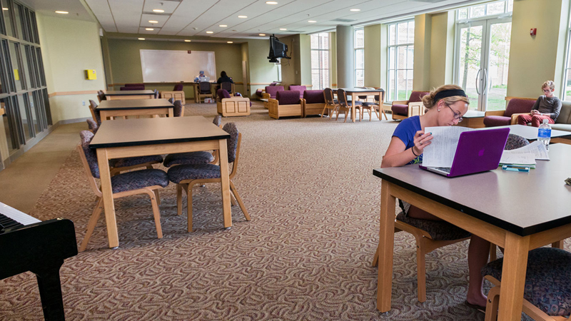 Students sit at different tables and chairs throughout the lounge space participating in different activities. There is a dry erase board mounted on the back wall.