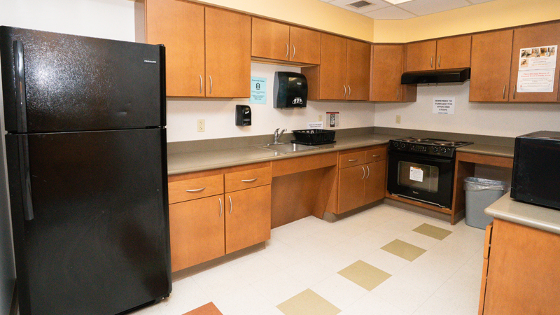 Counter space around the room in a U-shape, with a refrigerator in the back left corner, sink in the countertop with a paper towel dispenser mounted to the wall above the sink. There is an oven and stove along the back wall and a microwave is visible on the right side of the picture.