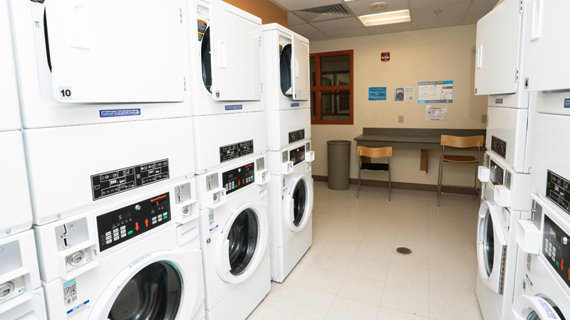 Stacked washer/dryer units on either side of the picture, with the washers on the bottom and dryers on the top. There is a folding table with a couple chairs along the back wall.