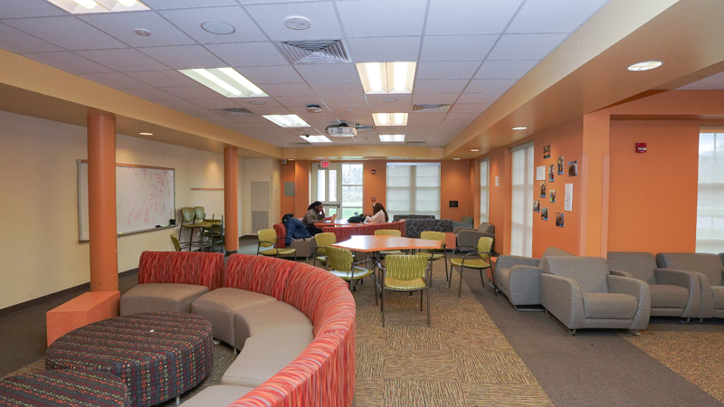 Lounge space with various different tables and chairs around the room. A dry erase board is mounted on the left wall, and two students sit at a table toward the back of the room. 