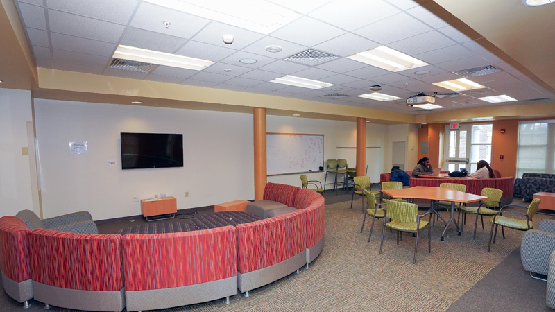 Lounge space with various different tables and chairs around the room. Lounge chairs are set up in a semi-circle around a TV mounted on the wall, and there is a dry erase board mounted farther back on the same wall. Two students sit at a table towards the back of the room.