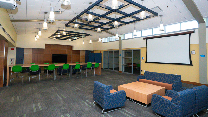 Lounge space with various different tables and seating options around the room. There is a TV mounted along the back wall and a large projector screen along the right wall, with entry to a smaller conference room.