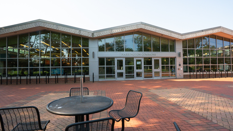 The front of the Harrington Commons building with ground to ceiling windows, bike racks on either side of the main entrance doors, and a table with four chairs at the front of the picture.  A brick walkway goes across the entire front of the building.