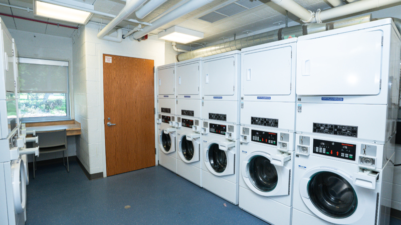 Five units of stacked washers and dryers with the washing machines on the bottom and dryers on the top. There are additional units along the left side of the picture, and a table with chair along the back wall in the room.