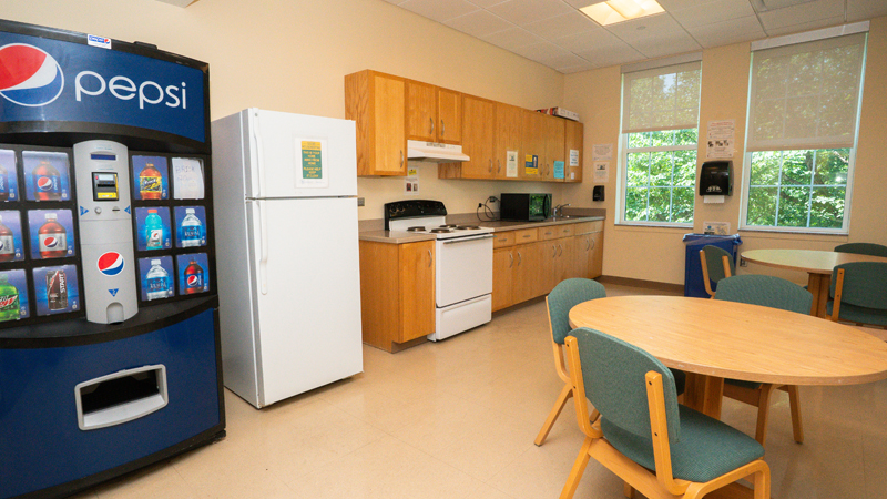 Along the left wall are a vending machine and refrigerator, and then cabinets along the second half of the wall. The oven and stove is built into the cabinets, and there is a microwave on top of the counterspace before the sink at the end. There are two window along the back wall with a mounted paper towel dispenser in between, and a recycling bin sits below. There are two round tables with chairs along the right side of the room. 