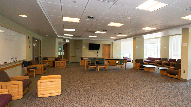 Lounge space with various different tables and chairs around the room. There is a dry erase board mounted to the left wall and a TV mounted on the back wall. There are windows along the right wall.