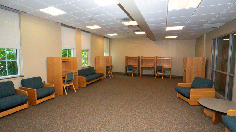 Lounge space with some lounge chairs and couches around the room, along with enclosed individual desks with chairs. 