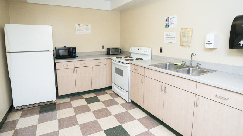 Picture of kitchen with refrigerator in back left corner, microwave and toaster oven on top of counter along the back wall. An oven and stove installed along the right side, next to a double sink. Many cabinets are below the counter space.