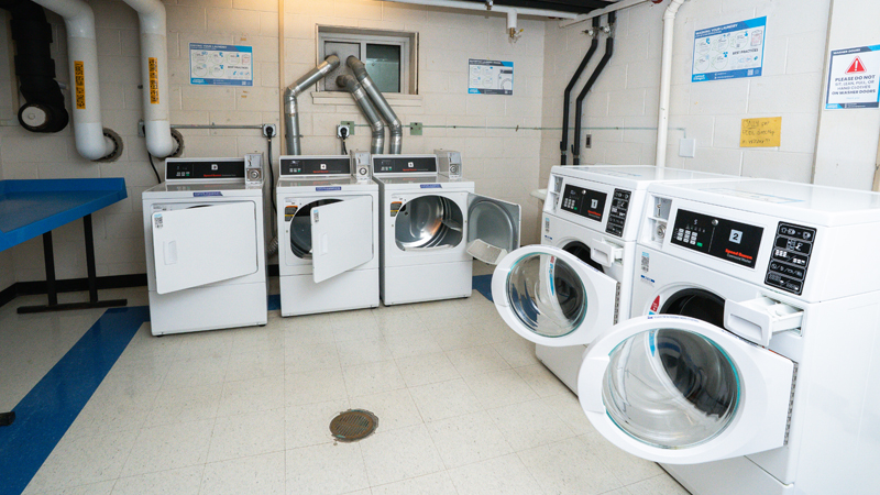 Picture of laundry room with three dryers along the back wall and two washers along the right wall. There is a table along the left wall.