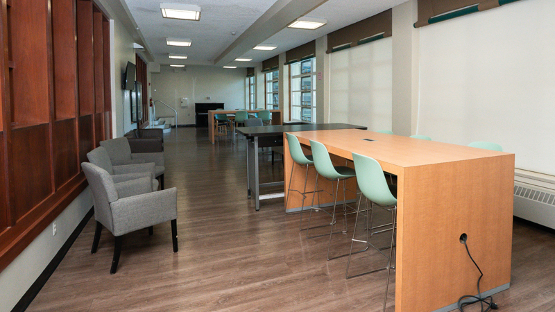 Lounge space with chairs along the left wall and various high-top tables and chairs along the middle of the space. Five large windows along the right side wall. 