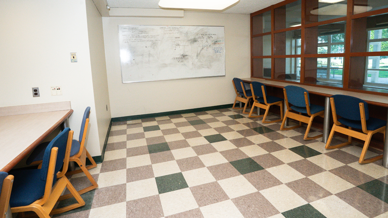 Lounge space with table tops along the left and right walls with chairs, and a dry erase board up on the back wall. 