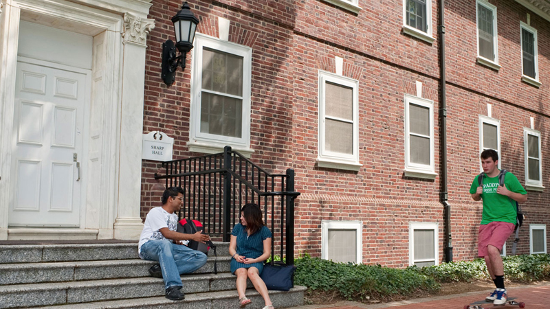 Two students sit on the steps leading up to Sharp Residence Hall, with other students passing by on the brick walkway in front of the building.
