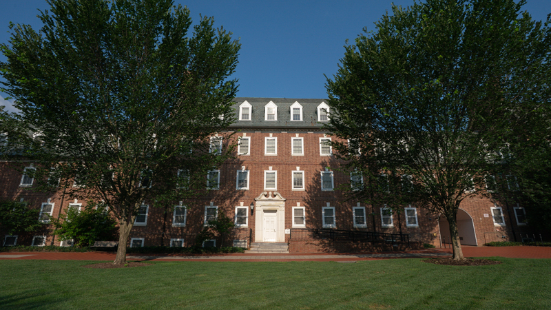 Straight ahead view of Sypherd Residence hall with main entrance door in the middle of the building and many windows visible across the front. Two large trees sit in a green field in front of a brick walkway in front of the building.