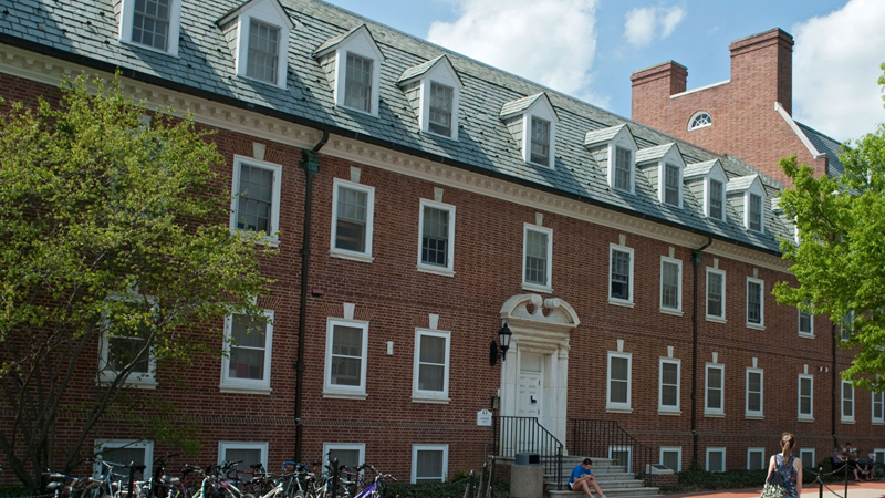 View of Sypherd Residence Hall with brick walkway along the front of the building, a bike rack to the left of the main door, and steps up to the entrance door.