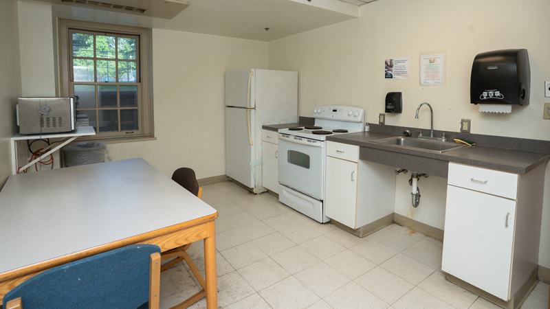 Along the right side of the wall are some cabinets and counter space with a sink, stove and oven unit, and refrigerator. A table with two chairs is in front of a mounted microwave on the left side of the room, with a window on the back wall.