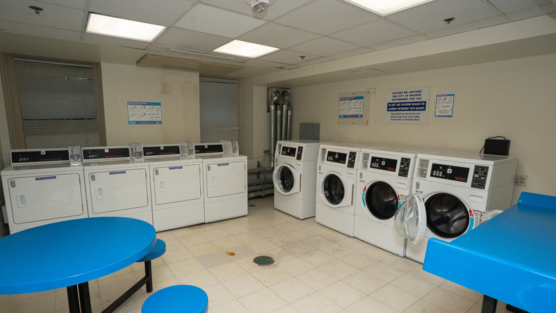 Four washing machines along the right wall and four dryers along the back wall, with a round table and seats and another high top table towards the front part of the room.