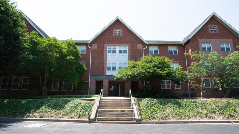 Steps lead up to a concrete walkway to the main entrance of Ray A Residence Hall. There are many trees in front of the building.