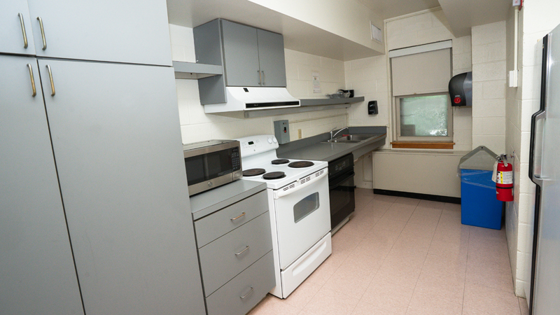 Along the left wall are cabinets with a stove and oven unit, an addiitonal oven unit, microwave on top of the country and a sink at the end of the counter space. There is a refrigerator along the right wall with a fire extinguisher mounted on the wall after it, before a recycling can in the back corner. There is a paper towel dispenser mounted on the right wall in the back of the space.