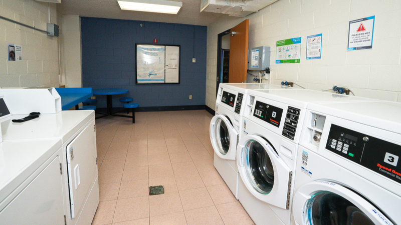 Washing machines on the right of the laundry room with dryer units on the left. There is a folding table next to another table with seats after the dryers on the left wall. 