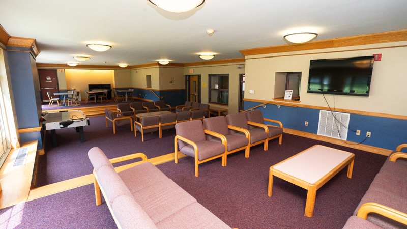 Lounge space with various different tables and chairs around the room. Both the front and back sections are raised compared to the main section when entering the space. There is a TV mounted on the wall in the front section of the space, an air hockey table in the middle, and a piano along the back wall.