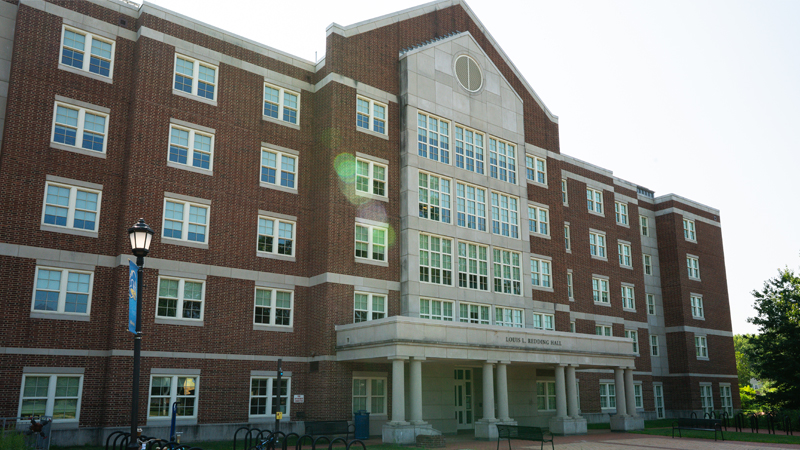 View of the front entranace of Louis Redding Residence Hall, with bike racks on either side of the main door and park benches along the brick walkway to the building entrance.