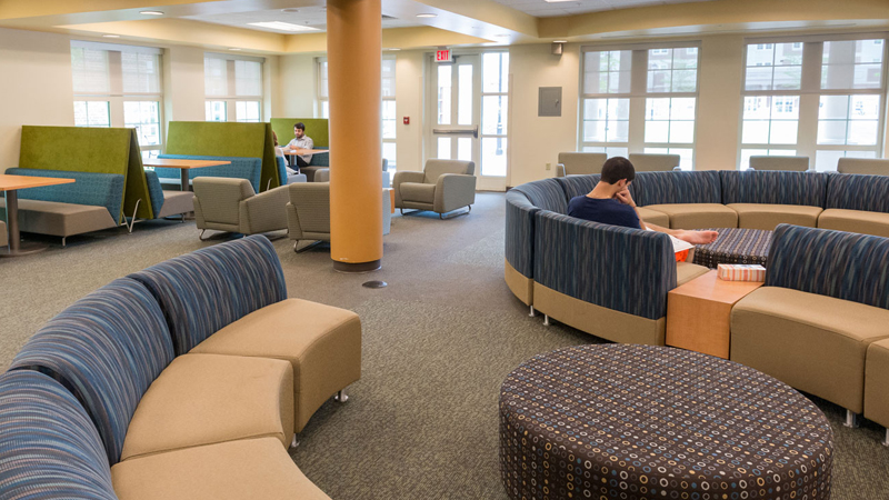 Students sitting in various chairs with different tables participating in different activities in the lounge space.