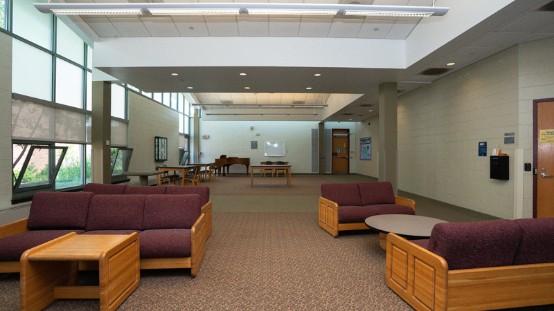 Lounge space with various different tables and chairs around the room. There is a piano in the back left corner and the door to the RA office on the right side of the picture with yellow signs on the door.