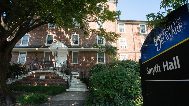 Outside view of main entrance to Smyth Residence Hall. Two sets of white stairs meet in the middle with another flight to the main entrance door. A large tree and other greenery surround the building. Sign of Smyth Hall in the right part of the picture.